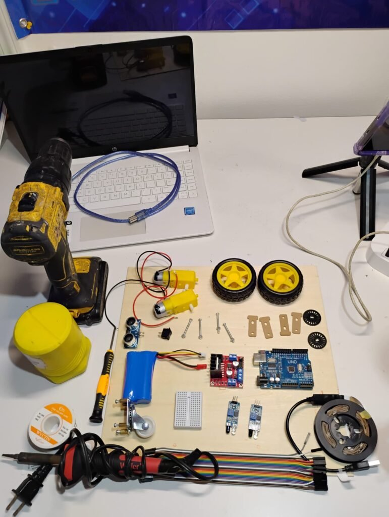 This image showcases a hands-on electronics and robotics setup featuring an Arduino Uno board, motor driver module, ultrasonic sensor, DC motors with wheels, rechargeable battery pack, soldering iron, breadboard, jumper wires, LED strip, and mechanical components ready for assembly. The workspace reflects a practical learning environment where students build, wire, program, and test real robotic systems.